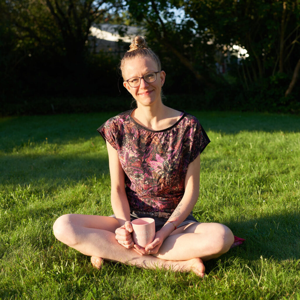 Lisa Jara sitting cross-legged on green grass in the morning sun, facing the camera, smiling, holding a pink cup of coffee saying "perfectly imperfect"